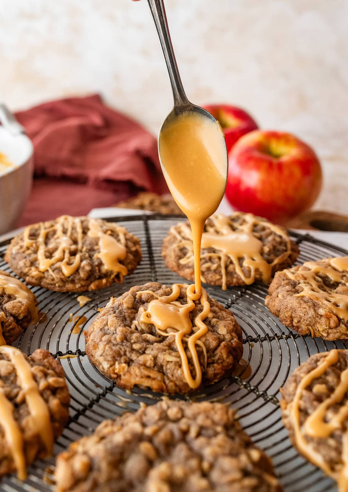 apple oatmeal cookies on cooling rack with salted caramel glaze being drizzled on top with a spoon.