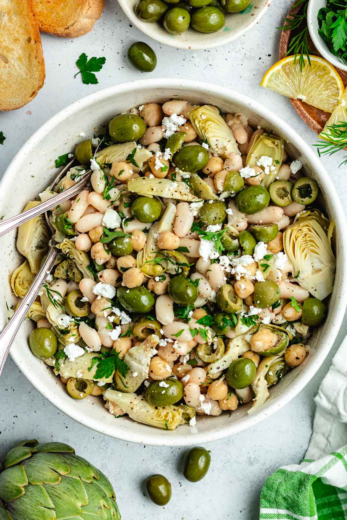 bean salad with artichokes, olives, and feat with herbs in bowl with serving spoons.