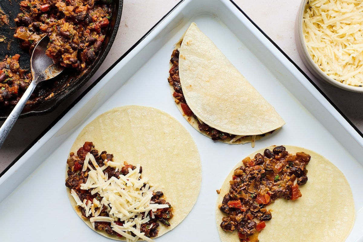 assembling crispy black bean tacos with black bean filling, tortillas, and cheese on sheet pan.