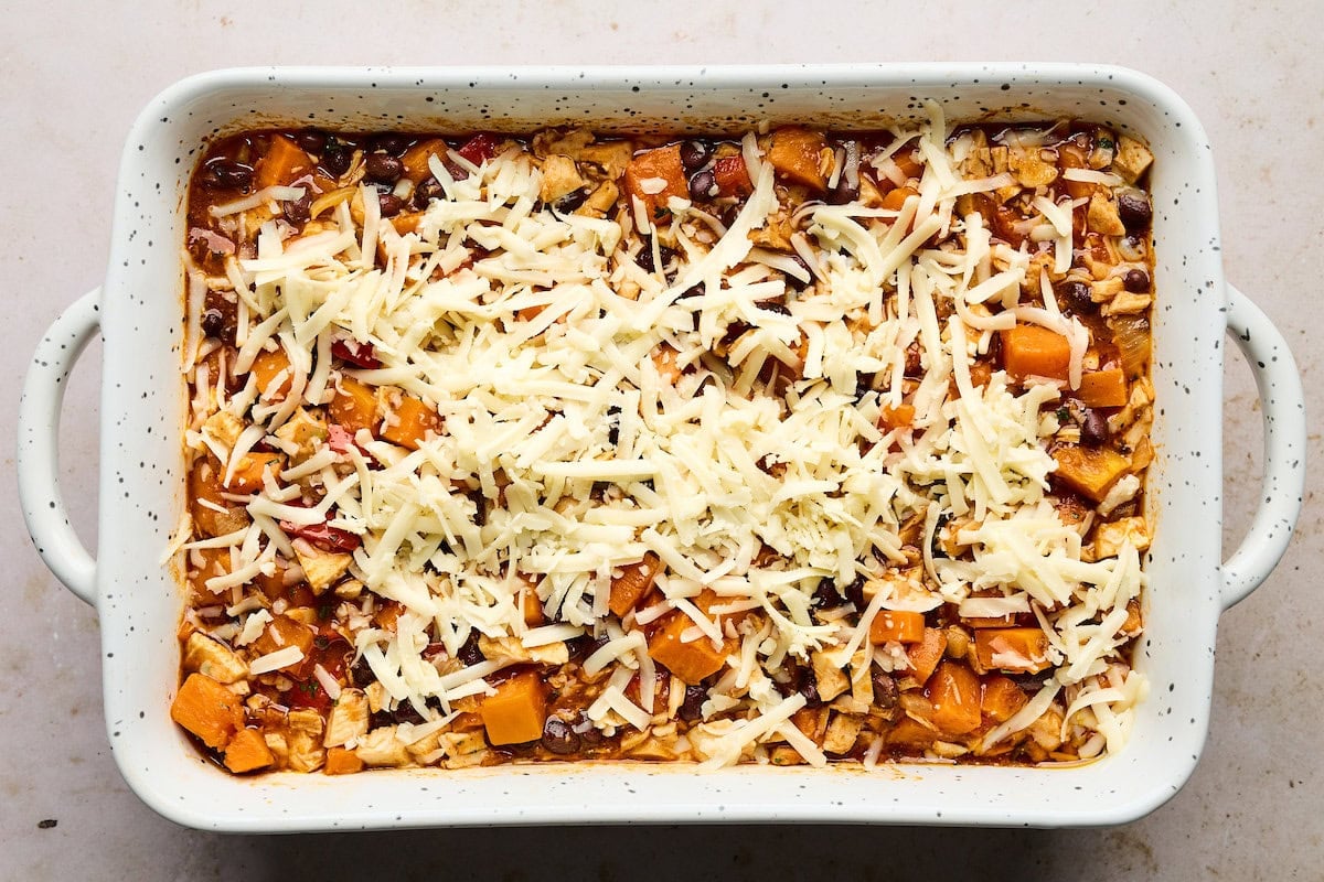 assembling sweet potato chicken enchilada bake in baking dish with shredded cheese on top.